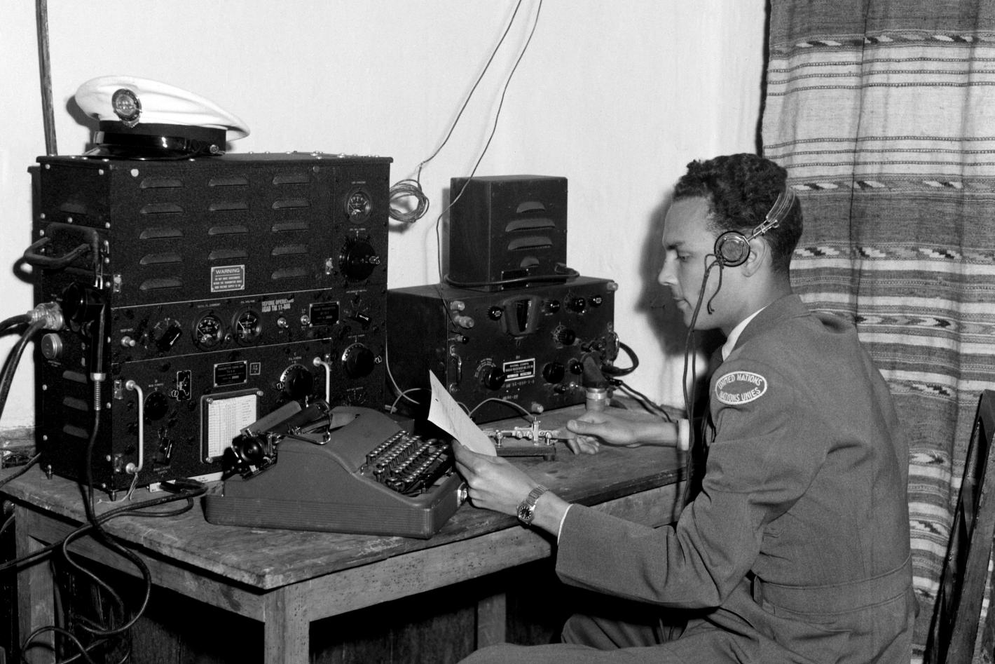 Black and white photograph of a young man in a military peacekeeping uniform siting at a desk with a headset on and a typewriter and other communication equipment in front of them.